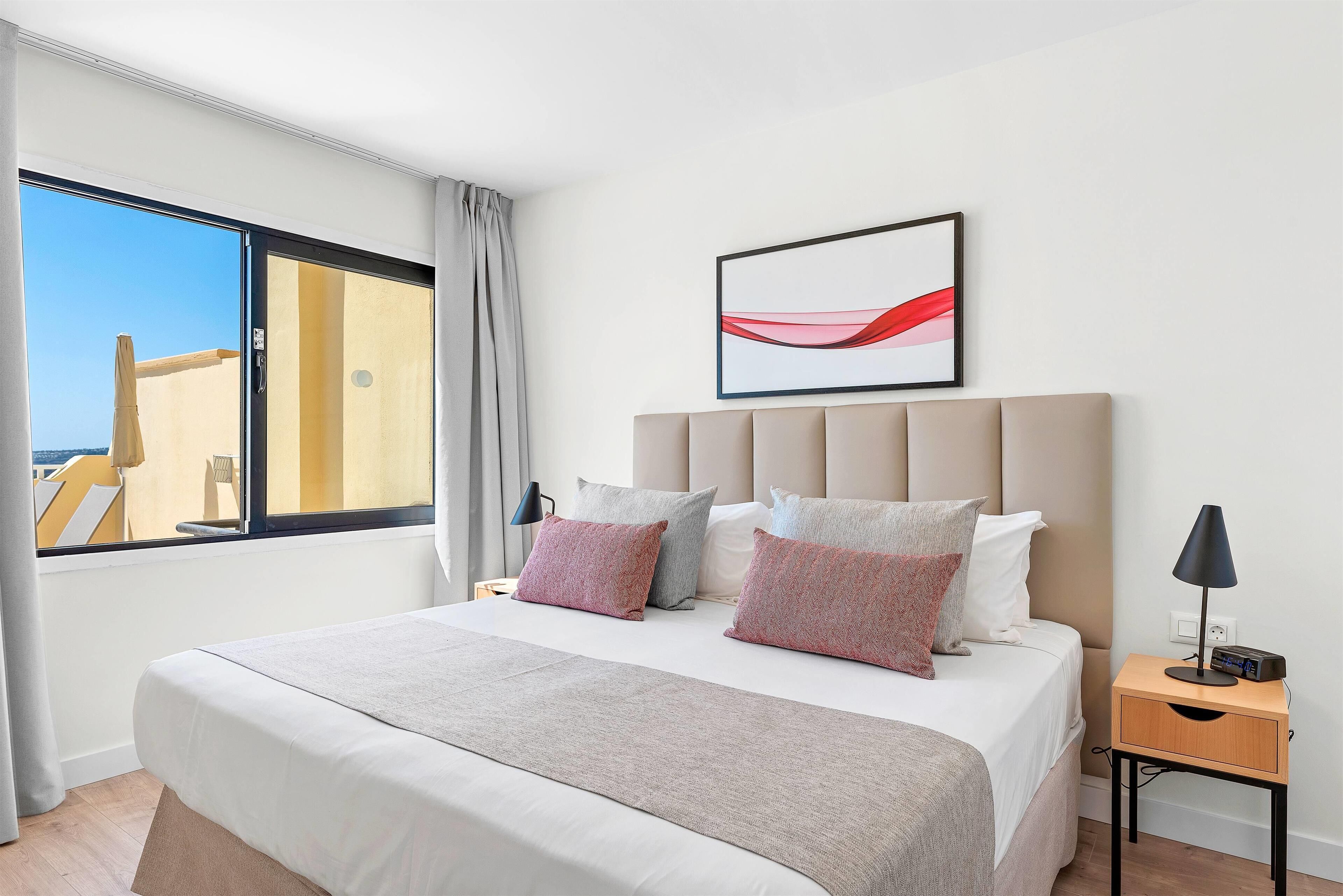 Ein modernes Hotelzimmer mit einem großen Bett, einem Nachttisch mit Lampe, einem gerahmten abstrakten Gemälde und Meerblick durch ein großes Fenster.