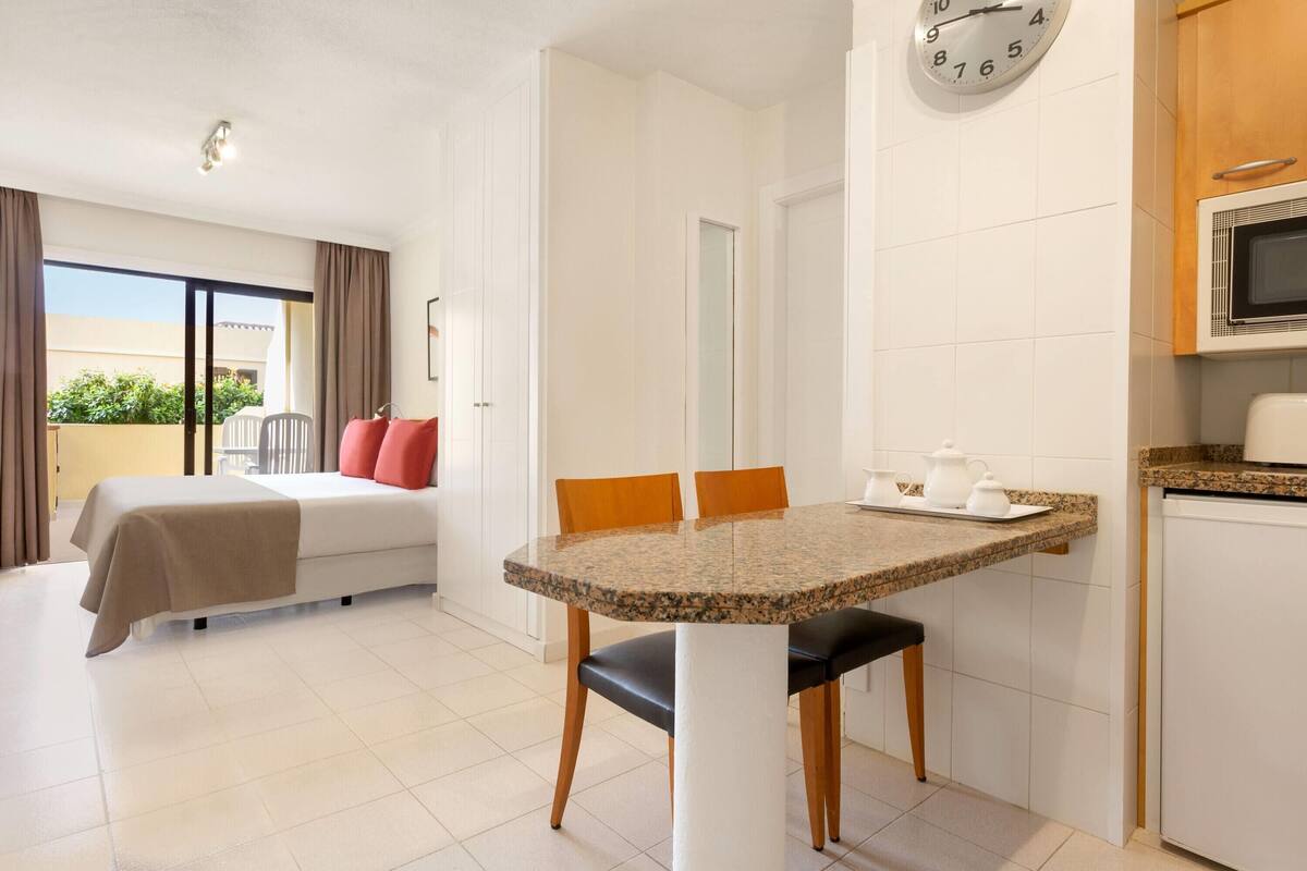 Ein Hotelzimmer mit einem Bett, einem Esstisch mit Stühlen, einer Mikrowelle und einer Uhr an der Wand.