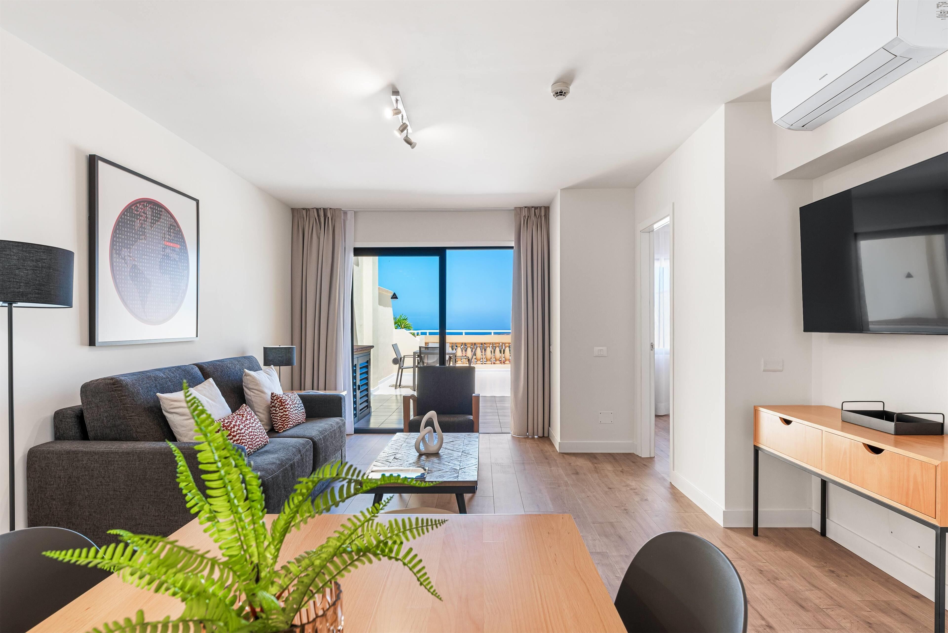 Una sala de estar moderna con sofá, mesa de centro y una mesa de comedor con sillas. Hay un televisor montado en la pared y un balcón con vistas al mar.