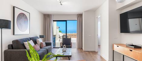 Una sala de estar moderna con sofá, mesa de centro y una mesa de comedor con sillas. Hay un televisor montado en la pared y un balcón con vistas al mar.