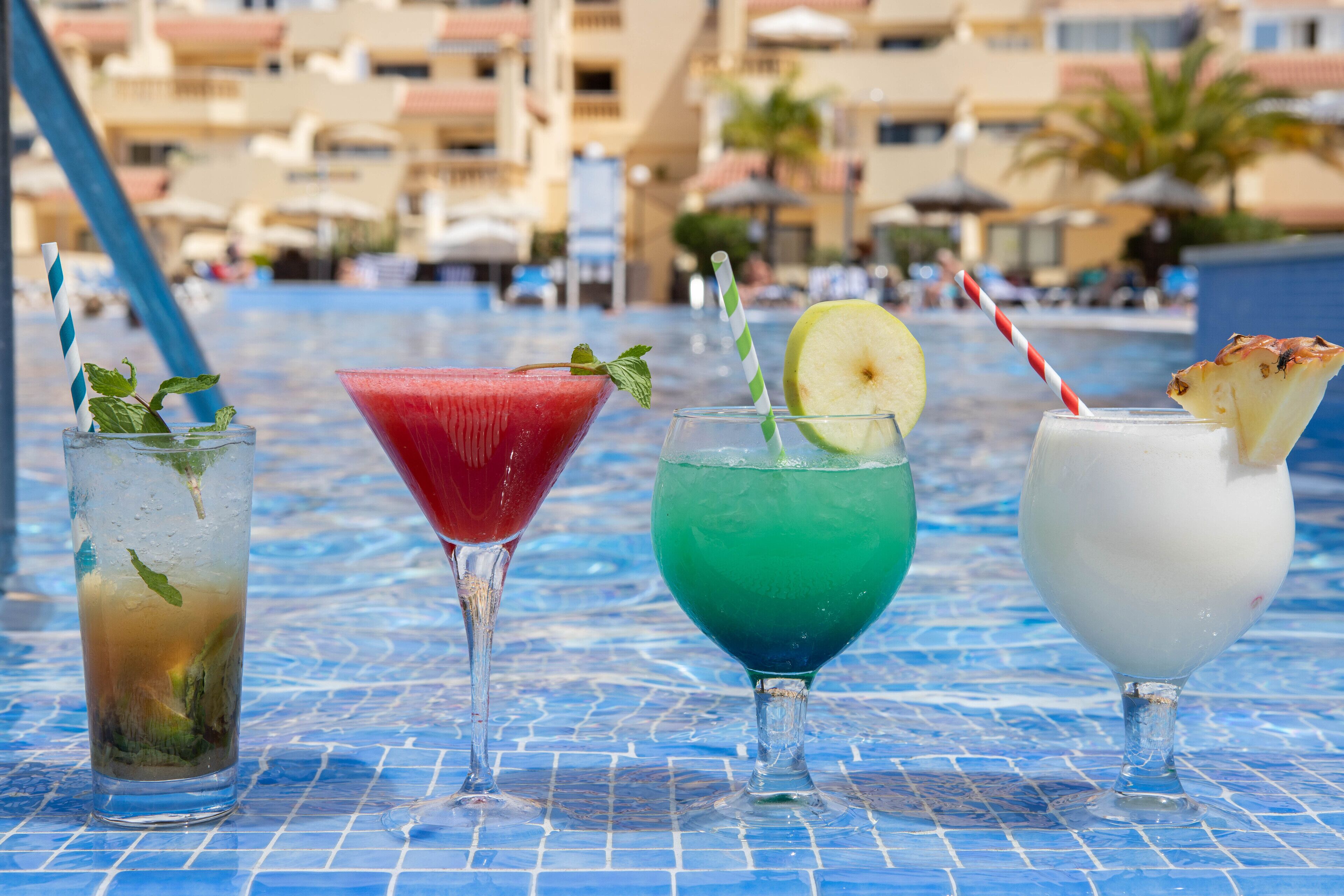 A poolside table with four drinks, a blue tiled pool, and a hotel background.