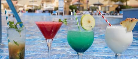 Ein Tisch am Pool mit vier Getränken, ein blau fliesiger Pool und ein Hotel im Hintergrund.