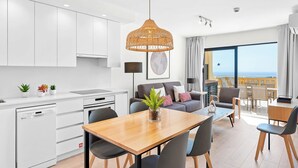 Une cuisine moderne avec des meubles blancs, un coin repas doté d’une table en bois et un salon avec un canapé et une vue sur la mer.