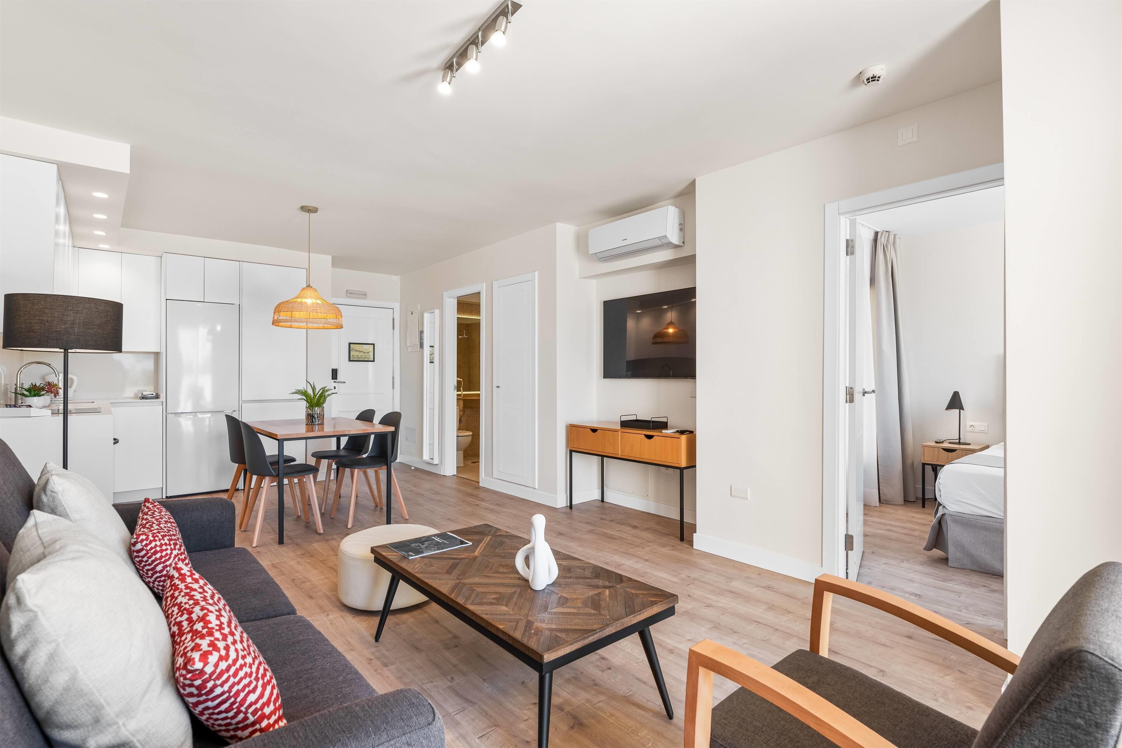 En moderne lejlighed med stue, spiseområde og køkken. Der er en sofa med puder, et spisebord med stole og en køkkenbordplade.
