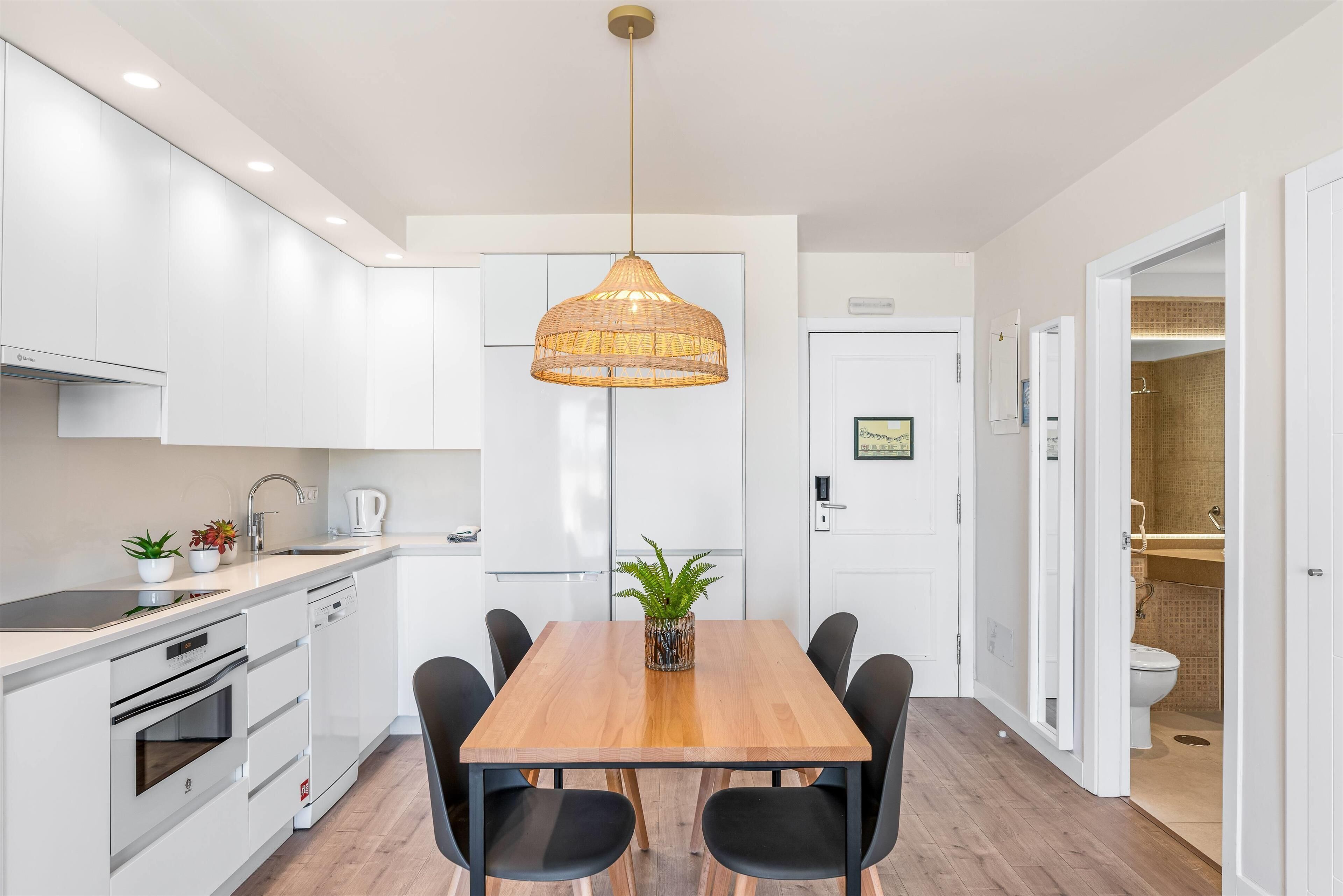 A modern kitchen with white cabinets, a wooden dining table, black chairs, and a hanging rattan light fixture.