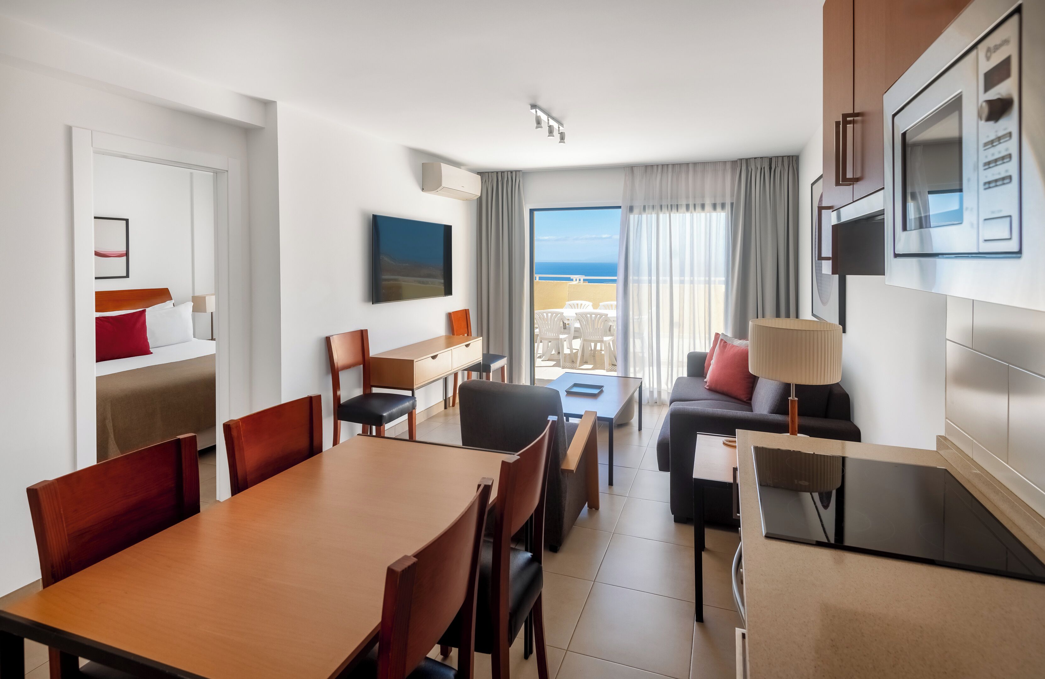 A hotel room with a dining area, a sofa, a TV, and a balcony with a view of the sea.