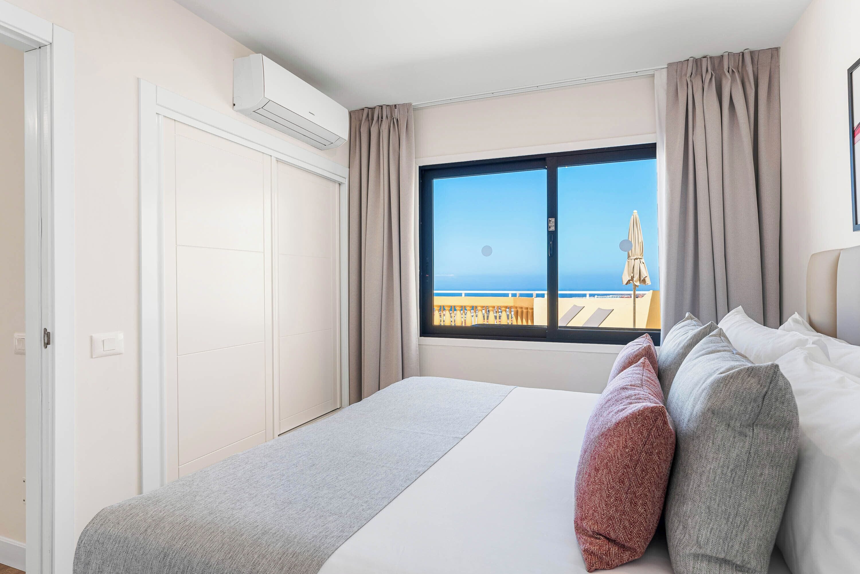 A hotel room with a bed, a window with a view of the sea, and an air conditioning unit.