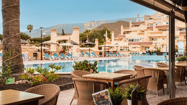 Un espace aménagé au bord de la piscine, avec des meubles en osier, une piscine et une vue sur un complexe hôtelier, avec des bâtiments et des montagnes en arrière-plan.
