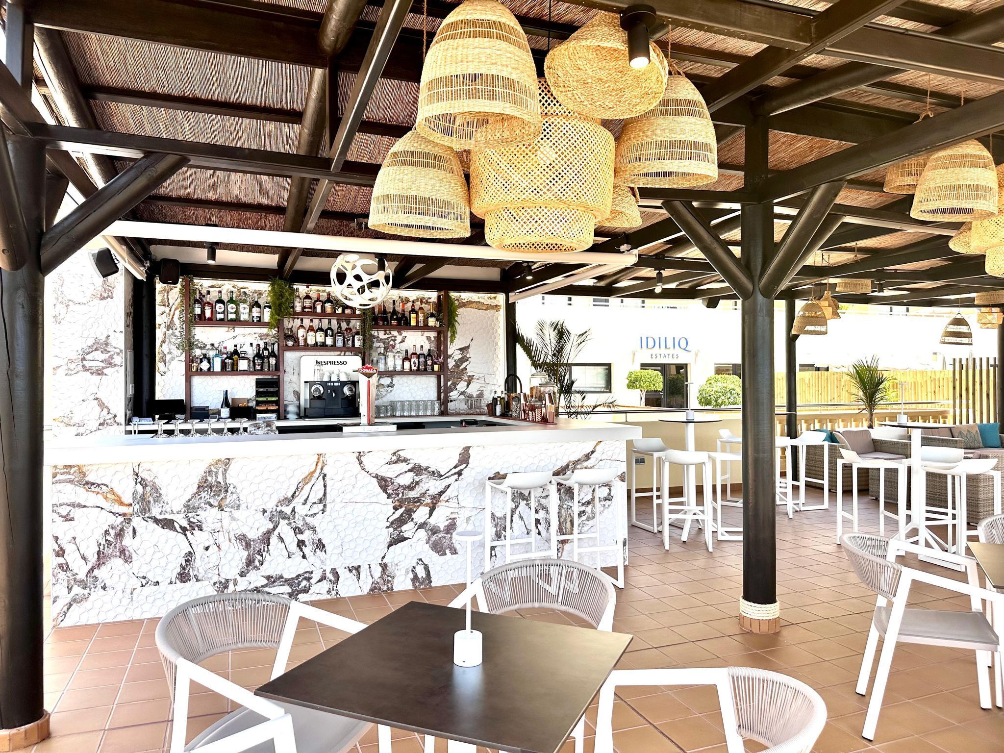 An outdoor bar area with wicker hanging lights, a bar counter with a Nespresso machine, and white wicker chairs.