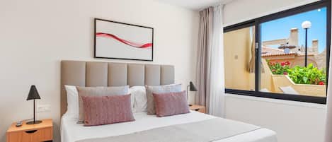 Habitación de hotel con cama, mesita de noche, lámpara, ventana con vista a un jardín y una obra de arte abstracto colgada en la pared.