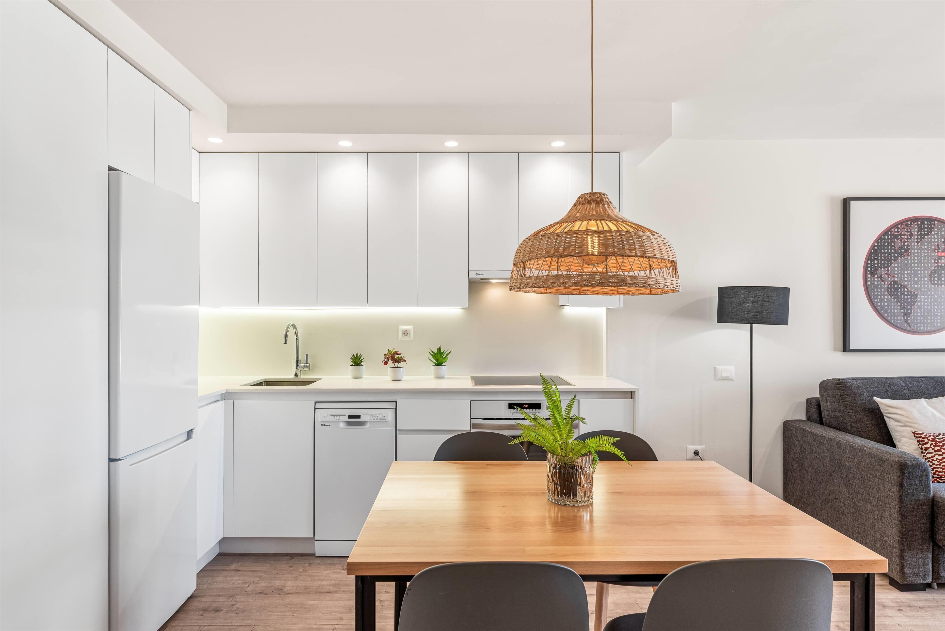 A modern kitchen with white cabinetry, a wooden dining table, and a hanging rattan light fixture.
