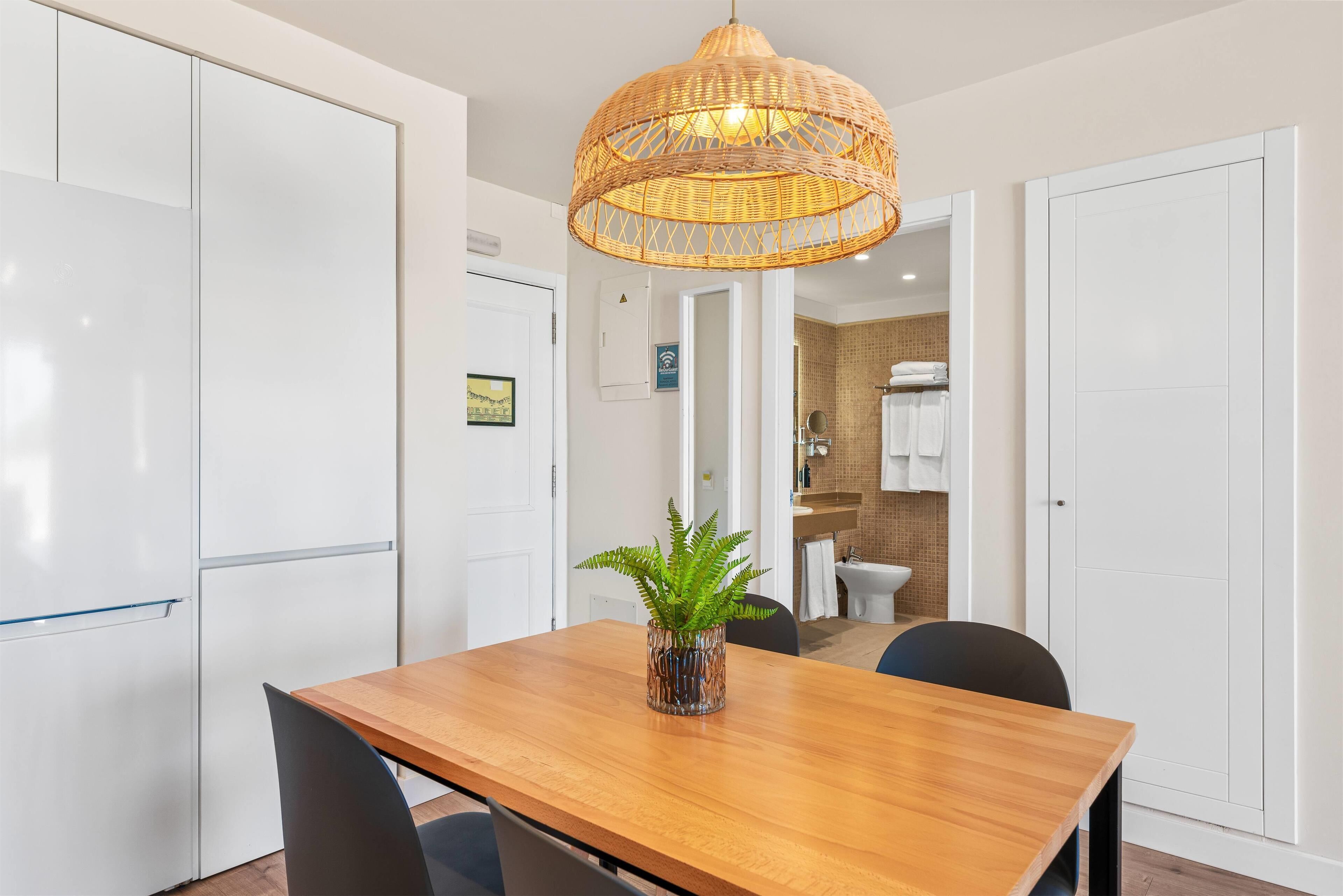 A modern dining area with a wooden table, chairs, a wicker pendant light, and a potted fern.