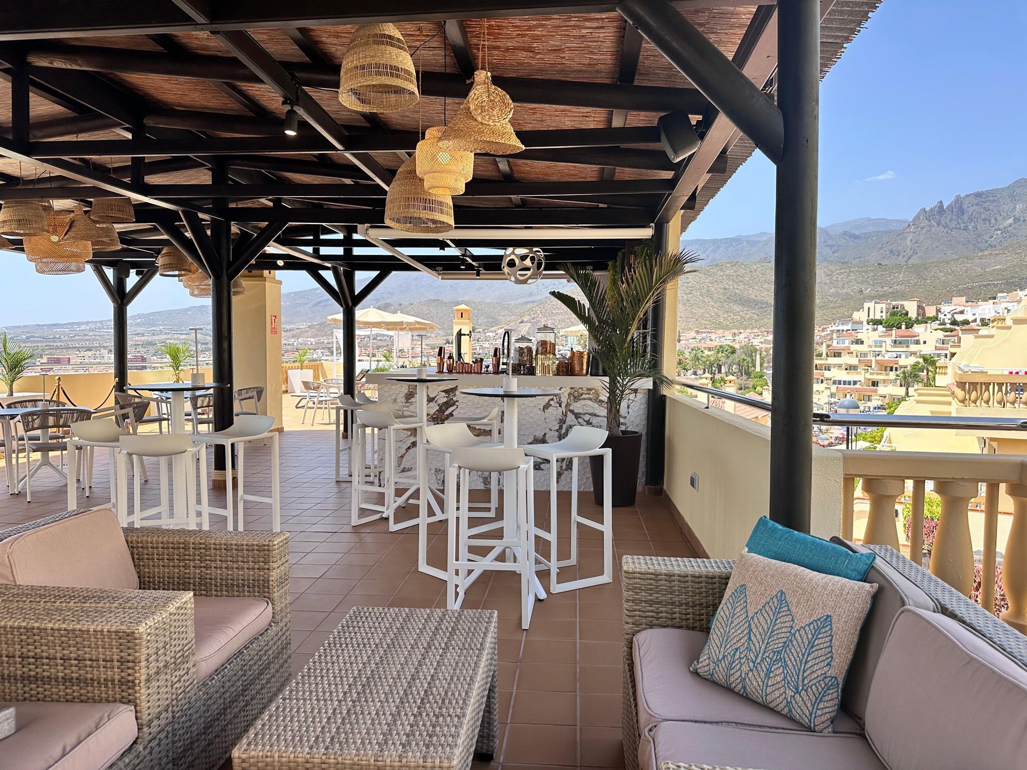 A covered outdoor seating area with wicker furniture and hanging lights, overlooking a mountainous landscape.
