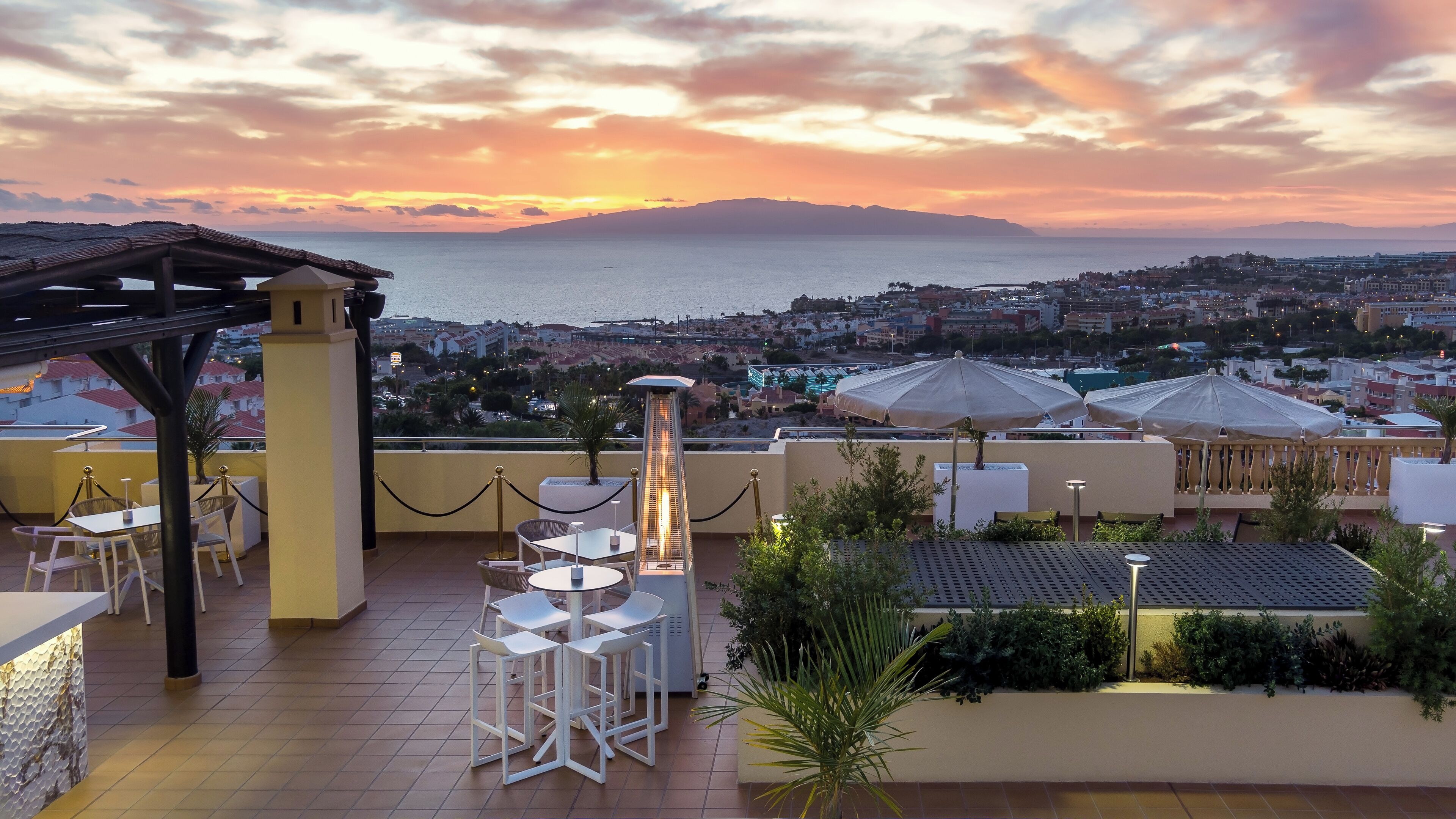 A rooftop terrace with tables and chairs, a decorative lamp, and a view of the sea and cityscape at sunset.