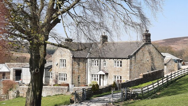 A stone building with multiple windows and a central entrance, surrounded by a stone wall and a grassy area. - Well appointed cottage on a working hill farm (Otley)