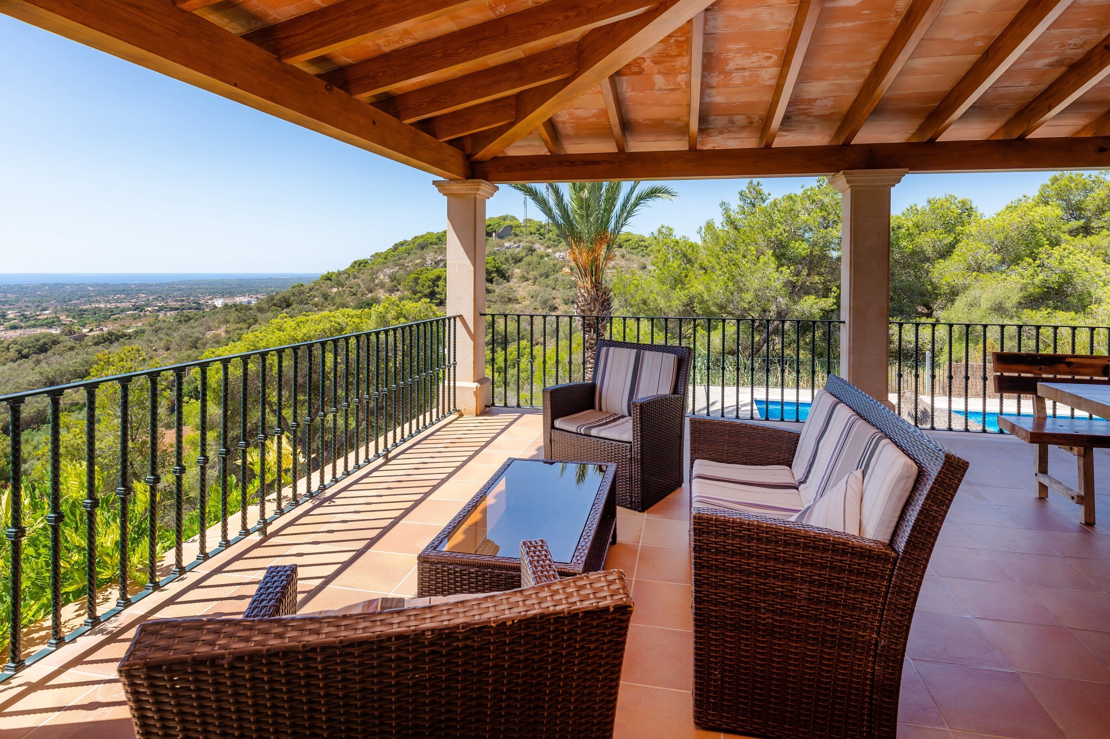 A covered outdoor seating area with wicker furniture, a glass-top table, and a view of the landscape.