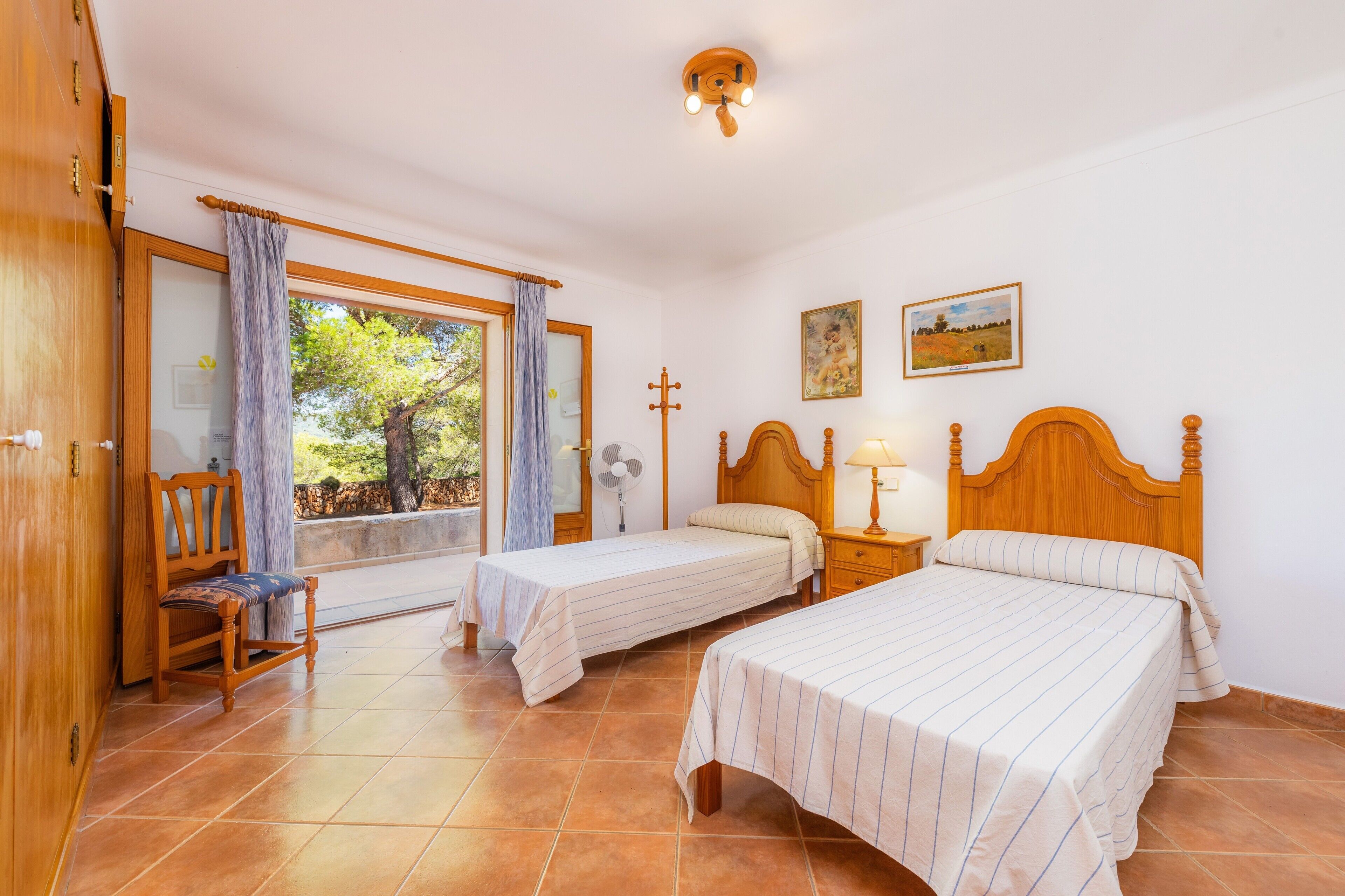 Un dormitorio con dos camas, una silla, una ventana con cortinas, un ventilador y una cruz en la pared.