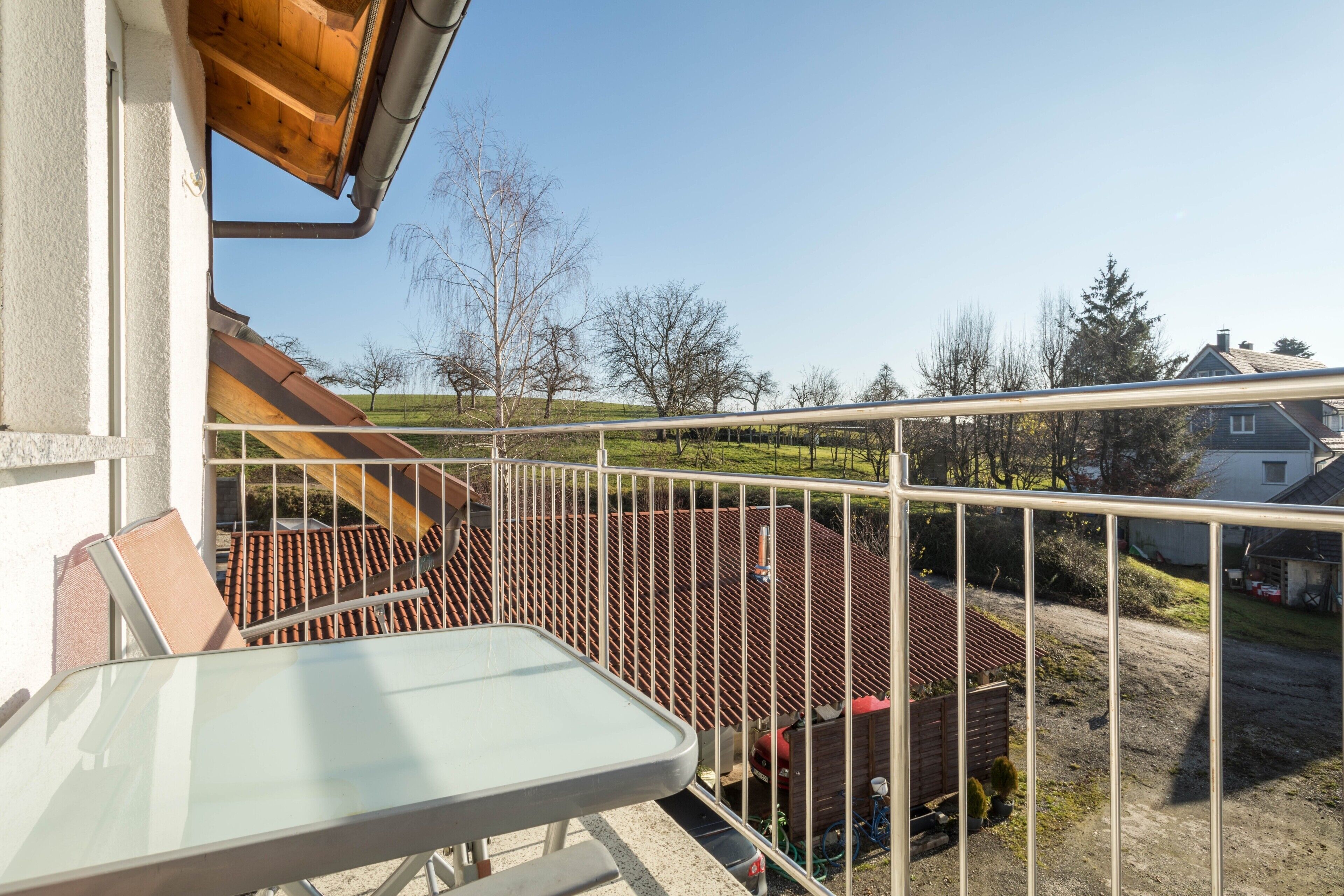 Un balcon doté d’une rambarde en métal, d’une table et offrant une vue sur une maison et des arbres.