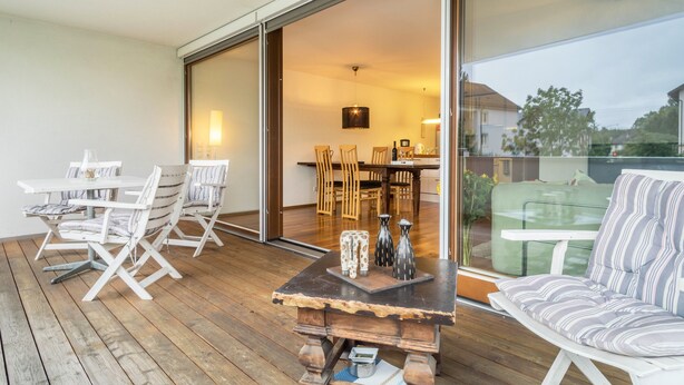 A wooden deck with a dining area, a sofa, and a kitchen visible through a sliding door. - Comfortable Holiday Apartment "Felix" with Balcony, Wi-Fi & TV (Bregenz)