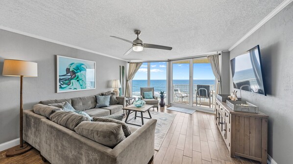 A spacious living room with a large gray sofa, a ceiling fan, and a view of the ocean through large windows. - Coastal Luxury - Shores Club Unit 604 (Daytona Beach Shores)