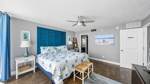 Un dormitorio con cama, ventilador de techo, mesita de noche, lámpara, silla, puerta y un cuadro en la pared.