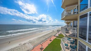 A beachfront hotel with a balcony overlooking the ocean, a paved walkway with outdoor seating, and a clear view of the beach and sea.