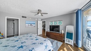 A bedroom with a bed, a ceiling fan, a dresser, a TV, and a view of the ocean through a window.