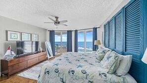 A bedroom with a large bed, a ceiling fan, a TV, and a view of the ocean through the window. - Coastal Luxury - Shores Club Unit 604 (Daytona Beach Shores)