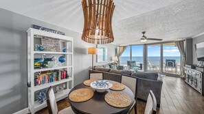 Una sala de estar moderna con comedor, que incluye una estantería, una mesa redonda con individuales tejidos, un ventilador de techo y vistas al océano.