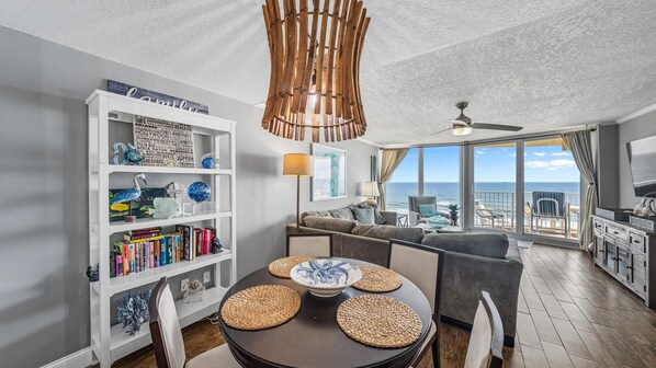 A modern living room with a dining area, featuring a bookshelf, a round table with woven placemats, a ceiling fan, and a view of the ocean.