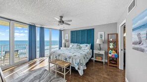 A bedroom with a large bed, a ceiling fan, a view of the ocean, a wooden floor, and a balcony with chairs. - Coastal Luxury - Shores Club Unit 604 (Daytona Beach Shores)
