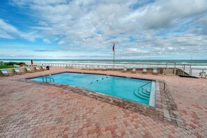 A swimming pool with a view of the ocean, surrounded by lounge chairs and a flag. - Coastal Luxury - Shores Club Unit 604 (Daytona Beach Shores)