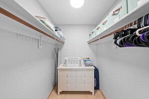 Un couloir étroit aux murs blancs, avec des poutres en bois et une armoire blanche dotée de tiroirs. Des étagères de part et d’autre contiennent divers objets, notamment des boîtes et des sacs.