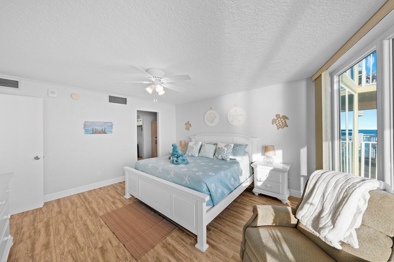 Quarto com cama branca, ventilador de teto, janela grande e vista para o oceano.