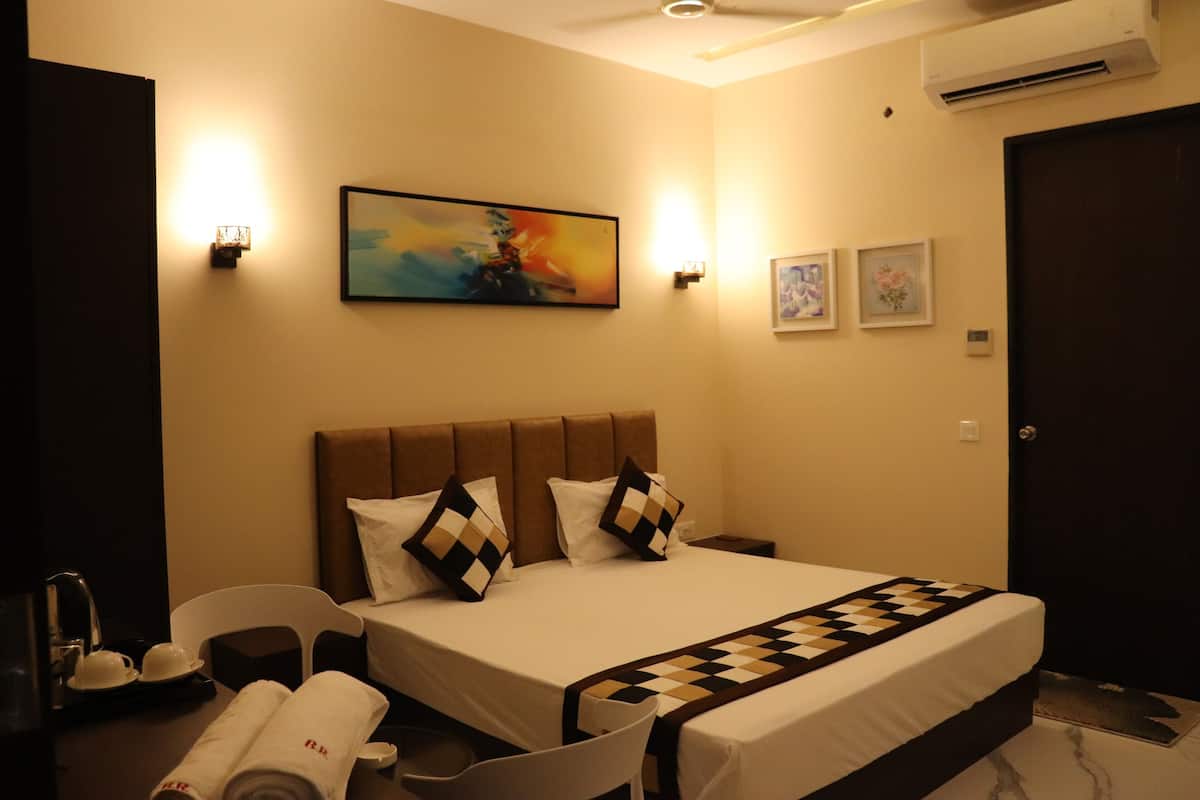 A hotel room with a large bed, a headboard, two bedside tables, a desk with a chair, a desk lamp, a wall-mounted air conditioner, and a painting on the wall.