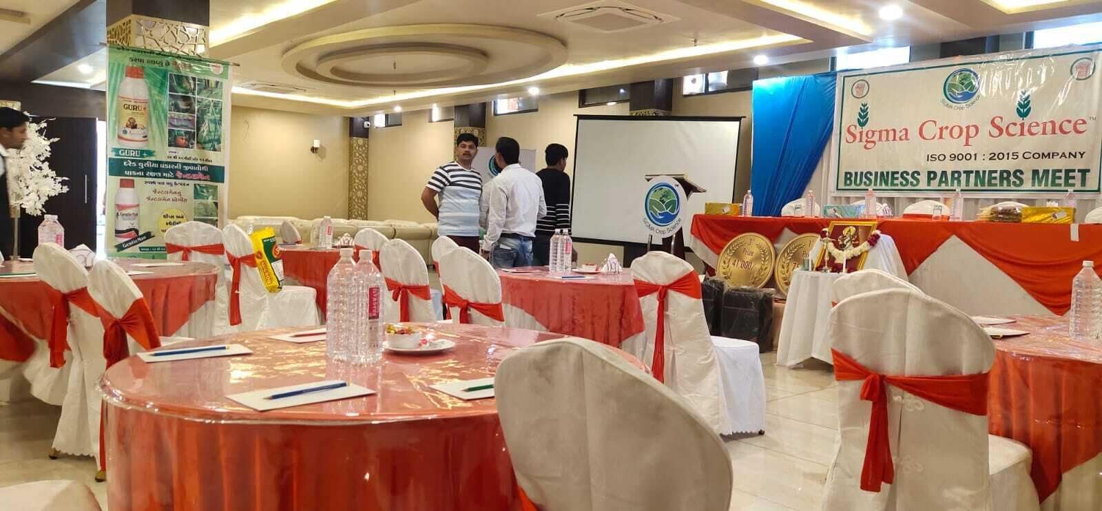 Un salón de conferencias con mesas redondas, sillas blancas con cintas rojas y un cartel para Sigma Crop Science.
