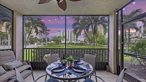 A balcony with a table set for two, featuring a view of palm trees and buildings. - Spacious Condo | Lanai, Pool and Steps to Beach (Marco Island)