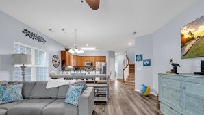A modern living room with a grey sofa, wooden flooring, and a dining area with wooden cabinets and a kitchen in the background. - Beautiful beach home! Private pool! Dates in August have been discounted! (Surfside Beach)