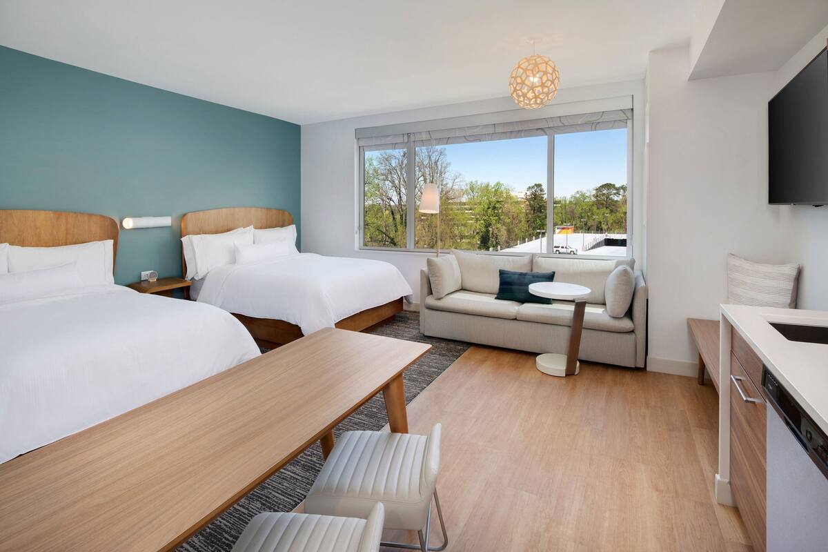 A modern hotel room with two beds, a sofa, a dining table, and a kitchen area.