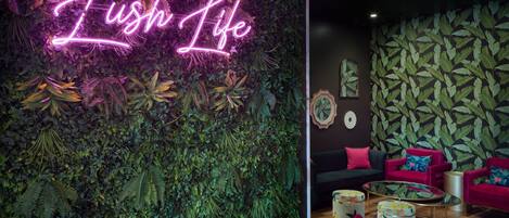 A room with a neon sign reading "Lush Life," a wall covered in green foliage, and a seating area with red chairs and a round table.