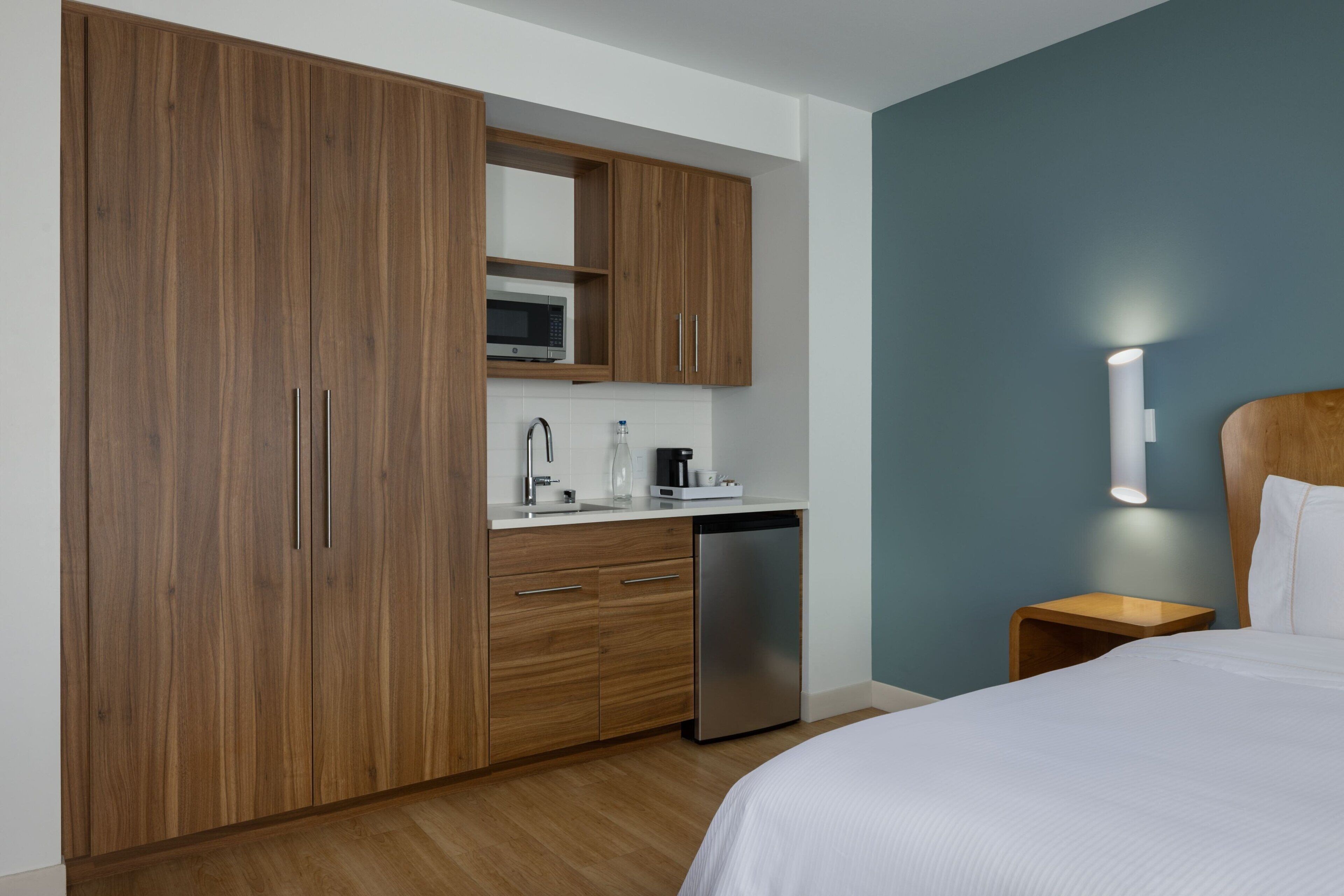 A modern hotel room with a wooden wardrobe, a built-in microwave, a sink, and a bedside table with a lamp.