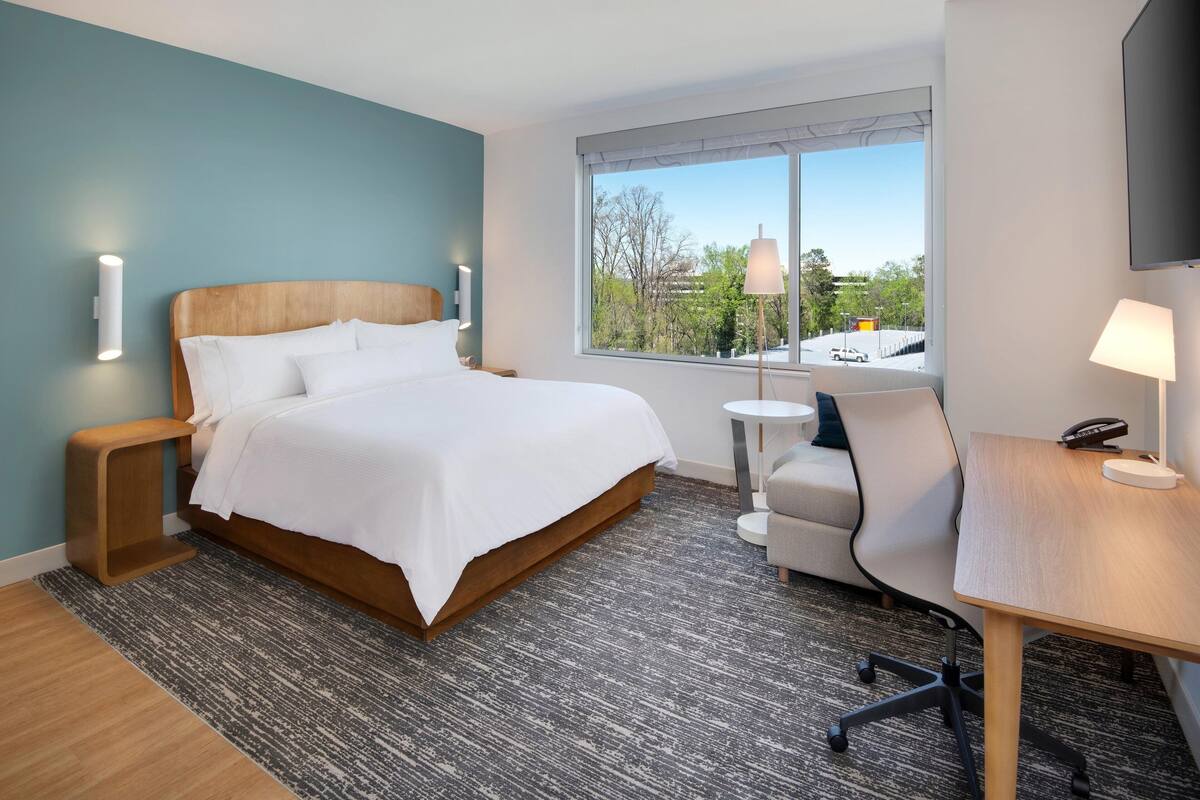 A modern hotel room with a large bed, a desk, a chair, a small table, and a TV.