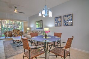 Uma área de refeições com mesa de vidro cercada por cadeiras de vime, ventilador de teto e luminárias fixadas na parede.