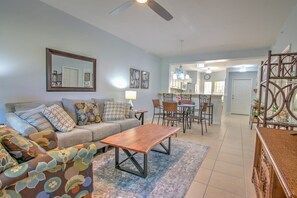 A living room with a sofa, coffee table, and patterned cushions. There is a dining area with chairs and a table, and a kitchen in the background.