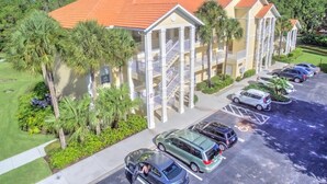 A multi-story hotel with a red-tiled roof, a parking lot with several cars, and palm trees in the foreground.