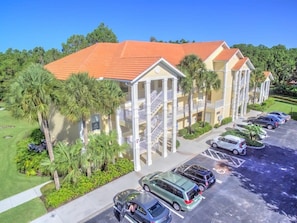 A multi-story hotel with a red-tiled roof, a parking lot with several cars, and palm trees in the foreground.