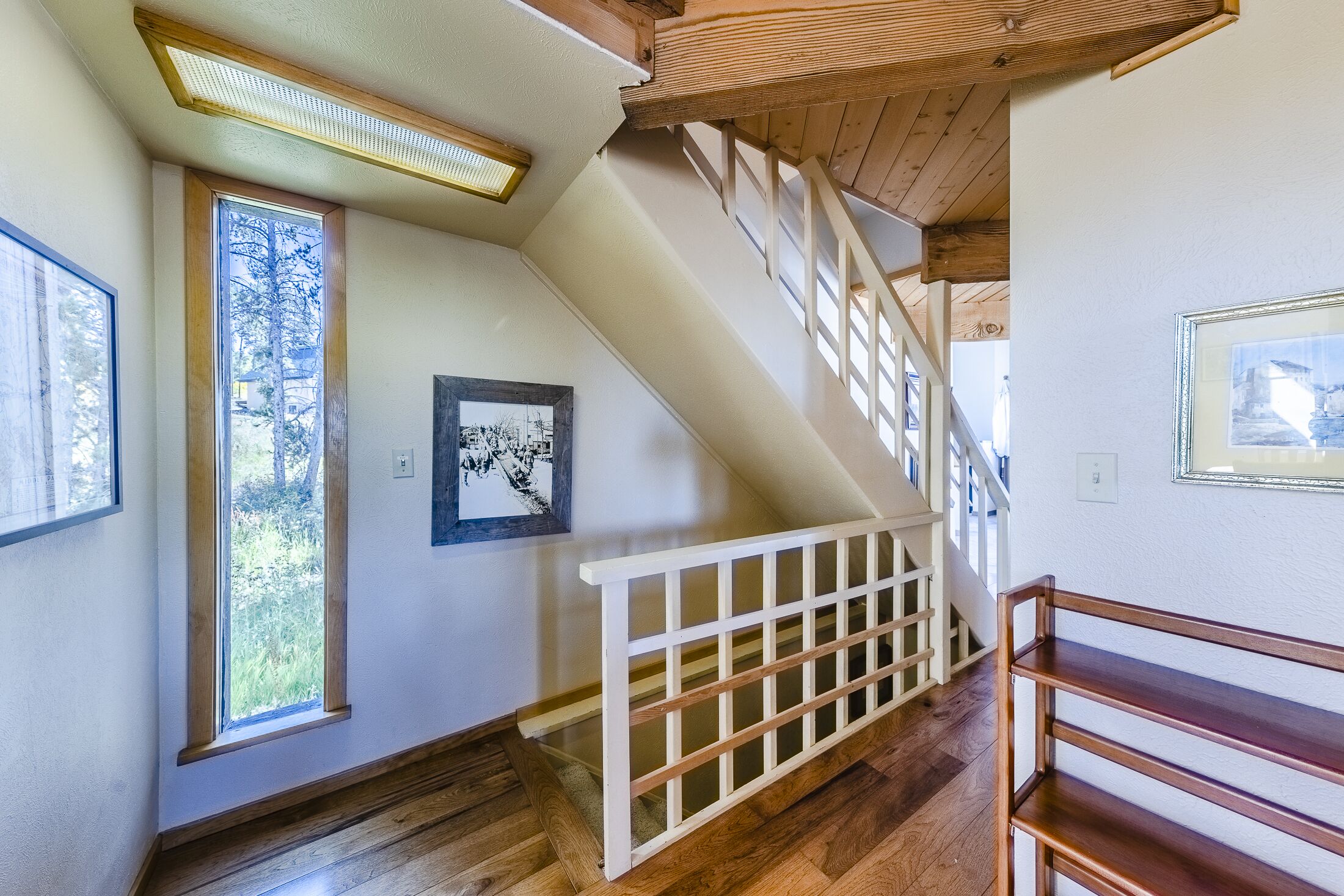 Een houten trap met een witte leuning, een groot raam met uitzicht op bomen en een lijst met een foto aan de muur.