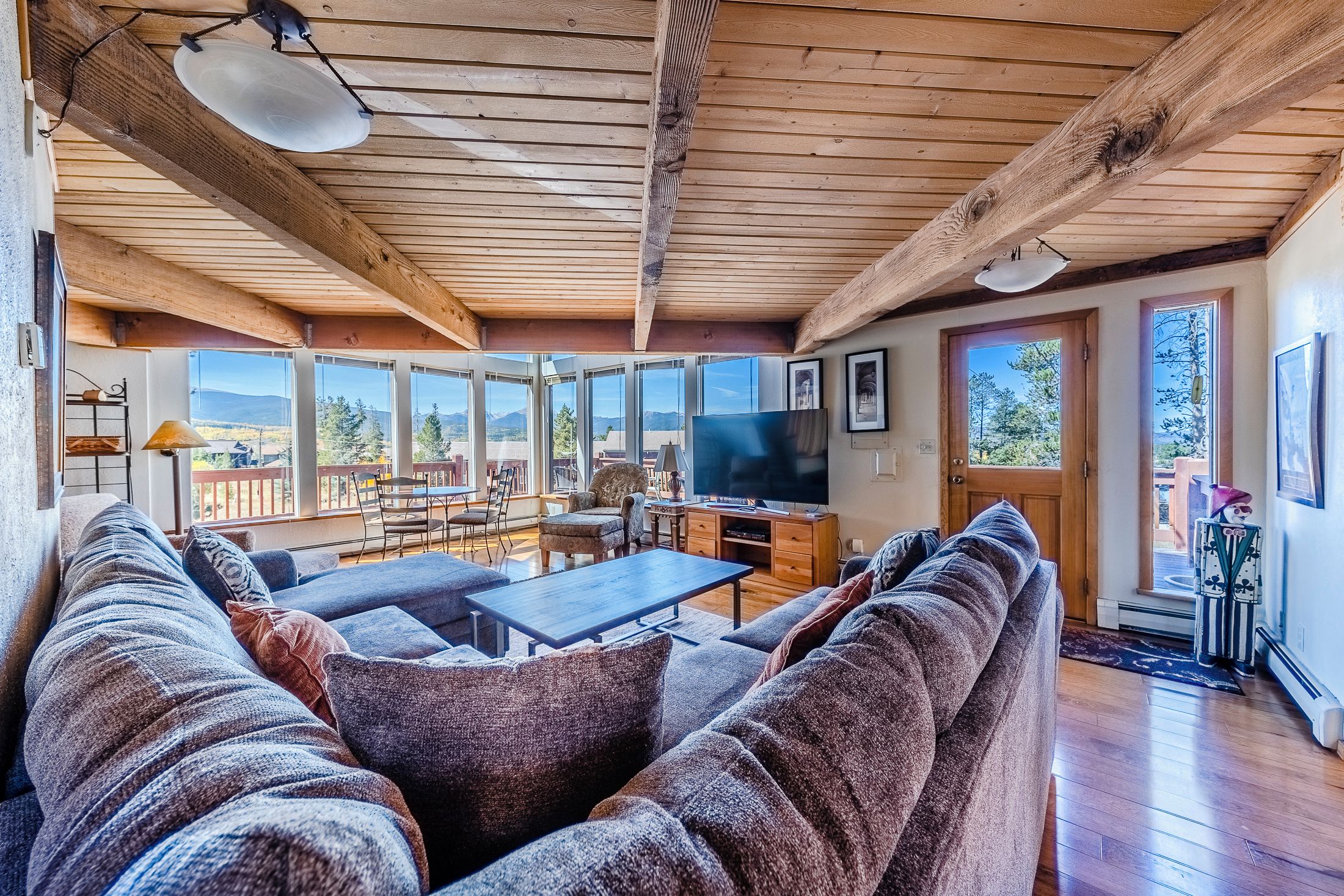 A cozy living room with a large sofa, a coffee table, and a TV. There are large windows offering a view of the outdoors.