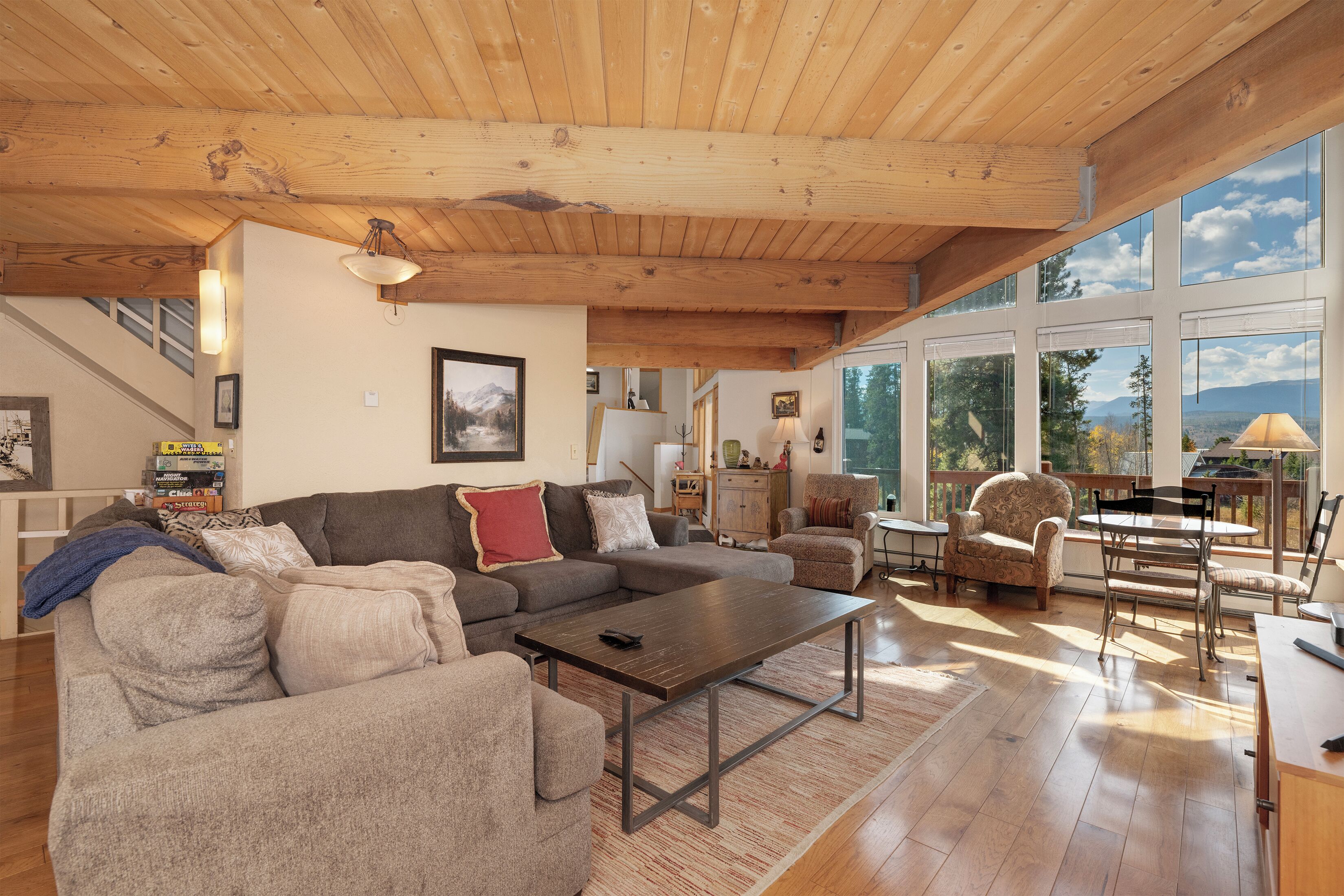 A spacious living room with a wooden ceiling, large windows, a sectional sofa, a coffee table, and a dining area with chairs and a table.