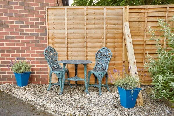 A patio with a wooden fence, two chairs, a small table, and two potted plants.